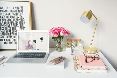 A study space that contains a laptop, lamp, notebooks, pair of glasses and a vase of pink roses. A picture is featured that contains positive messages about learning and strength.